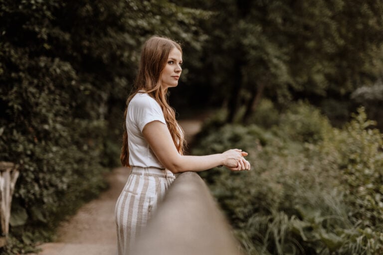 Eine junge Frau mit langen Haaren steht im Freien und lehnt sich an ein Holzgeländer. Sie trägt ein weißes T-Shirt und gestreifte Hosen. Der Hintergrund ist üppig und grün, durch die Bäume ist ein schmaler Pfad sichtbar. Sie blickt zur Seite zur Portraitfotografin aus Havixbeck.