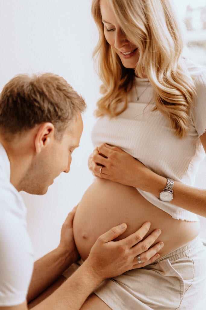 Eine schwangere Frau mit langen blonden Haaren lächelt auf ihren Babybauch herab, während ein Mann, ebenfalls blond, kniet und sanft ihren Bauch berührt. Beide tragen lässige weiße Kleidung und wirken glücklich und aufgeregt. Die Szene ist warm beleuchtet und mit natürlichem Licht gefüllt, perfekt für Babybauchfotografie von einer talentierten Familienfotografin.