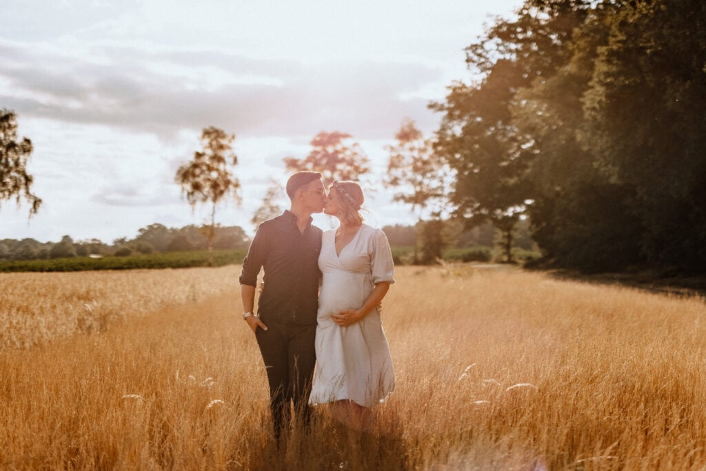 Ein Paar steht auf einer sonnenbeschienenen Wiese mit hohem Gras. Der Mann küsst die Stirn der Frau, während sie ihren schwangeren Bauch hält. Die Frau trägt ein weißes Kleid und der Mann ein dunkles Outfit. Im Hintergrund sind Bäume und ein bewölkter Himmel zu sehen, ein wunderschöner Moment der Babybauchfotografie in Havixbeck und Umgebung.