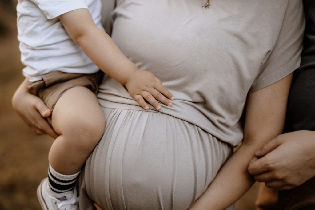 Eine schwangere Frau berührt zärtlich ihren Babybauch, während ein Kind in weißem Hemd und braunen Shorts seine Hand auf ihren Bauch legt. Eine weitere Person steht neben ihr und hat den Arm um ihre Taille gelegt. Das Bild, aufgenommen von der Familienfotografin in Havixbeck und Umgebung, vermittelt ein Gefühl von Familie und Vorfreude.