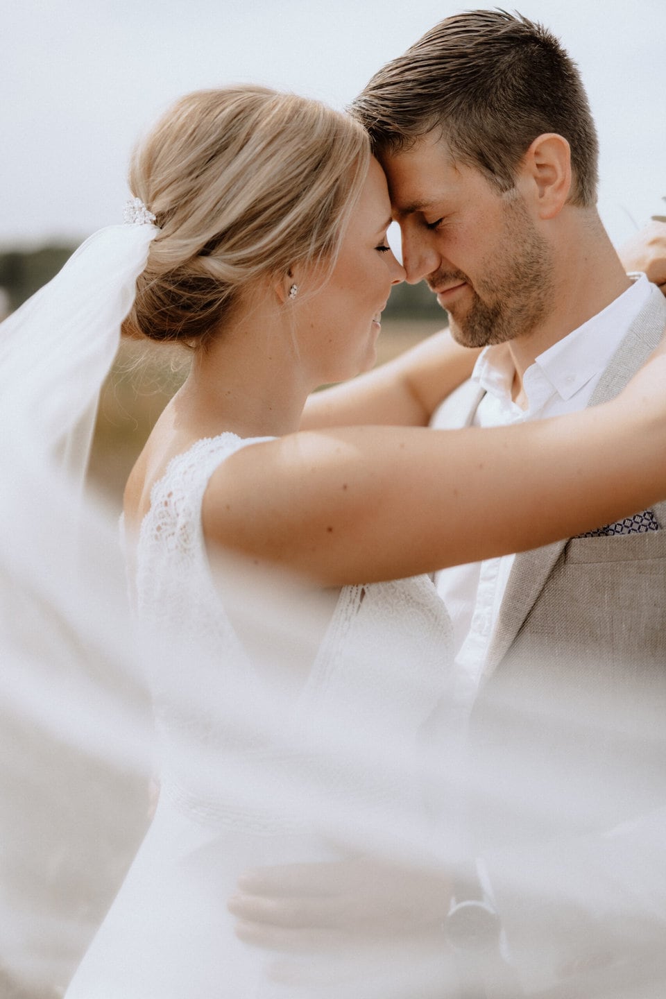 Braut und Bräutigam umarmen sich eng, ihre Stirnen berühren sich, ihre Augen sind geschlossen und sie wirken zufrieden. Die Braut trägt ein weißes Kleid mit Schleier und der Bräutigam einen hellen Anzug. Der Schleier weht um sie herum.