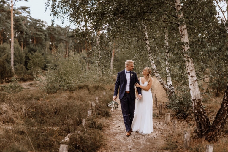 Braut und Bräutigam gehen Hand in Hand auf einem Feldweg durch ein Waldstück. Der Bräutigam trägt einen dunklen Anzug und eine Fliege, die Braut ein weißes Brautkleid und hält einen Blumenstrauß in der Hand. Bäume und Grün umgeben sie und schaffen eine heitere Atmosphäre.Einfühlsam aufgenommen von der Hochzeitsfotografin aus dem Münsterland.