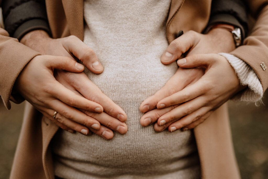 Nahaufnahme von zwei Paar Händen, die einen Babybauch sanft wiegen. Die oberen Hände gehören einer Person, die eine beige Jacke trägt, und die unteren Hände gehören einer anderen Person, die einen cremefarbenen Pullover trägt. Der Hintergrund ist leicht verschwommen und fängt die Essenz der Babybauchfotografie in Münster und Umgebung ein.