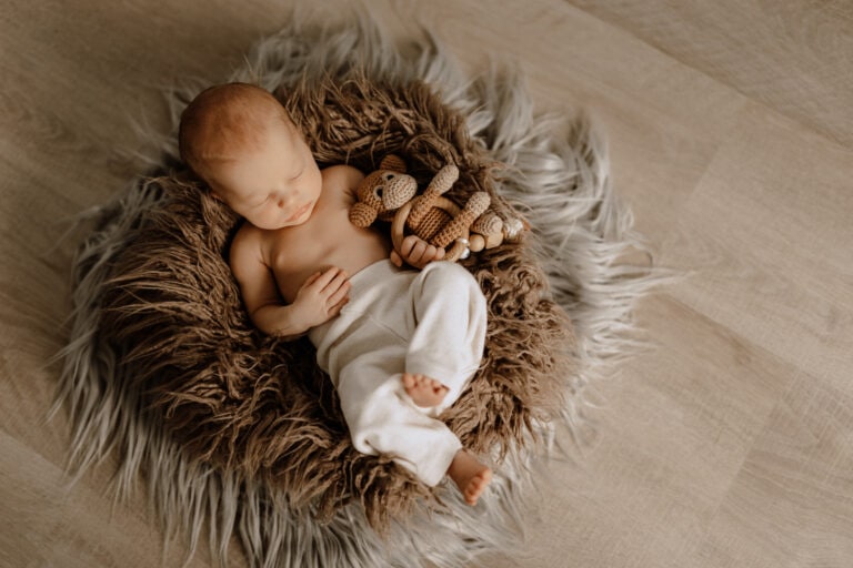 Ein Baby schläft friedlich auf dem Rücken, eingekuschelt in einem runden Bett aus flauschigem, braunem Fell auf einem Holzboden. Das Neugeborene trägt weiße Hosen und ist oben ohne, mit einem gehäkelten Tierspielzeug unter dem Arm. Dieser heitere Moment wurde vom Newborn Fotograf aus Billerbeck und Nottuln perfekt eingefangen.