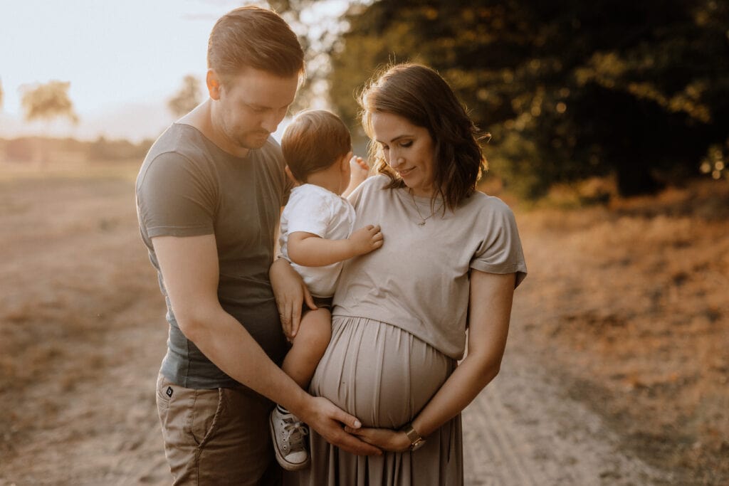 Ein Paar steht bei Sonnenuntergang draußen, der Mann links und die Frau rechts, die zwischen ihnen ein Kleinkind halten. Die Frau wiegt ihren Babybauch, während der Mann sanft seine Hand darauf legt. Sie alle blicken liebevoll auf ihren wachsenden Bauch, perfekt für eine Familienfotografin, um diesen kostbaren Moment festzuhalten.