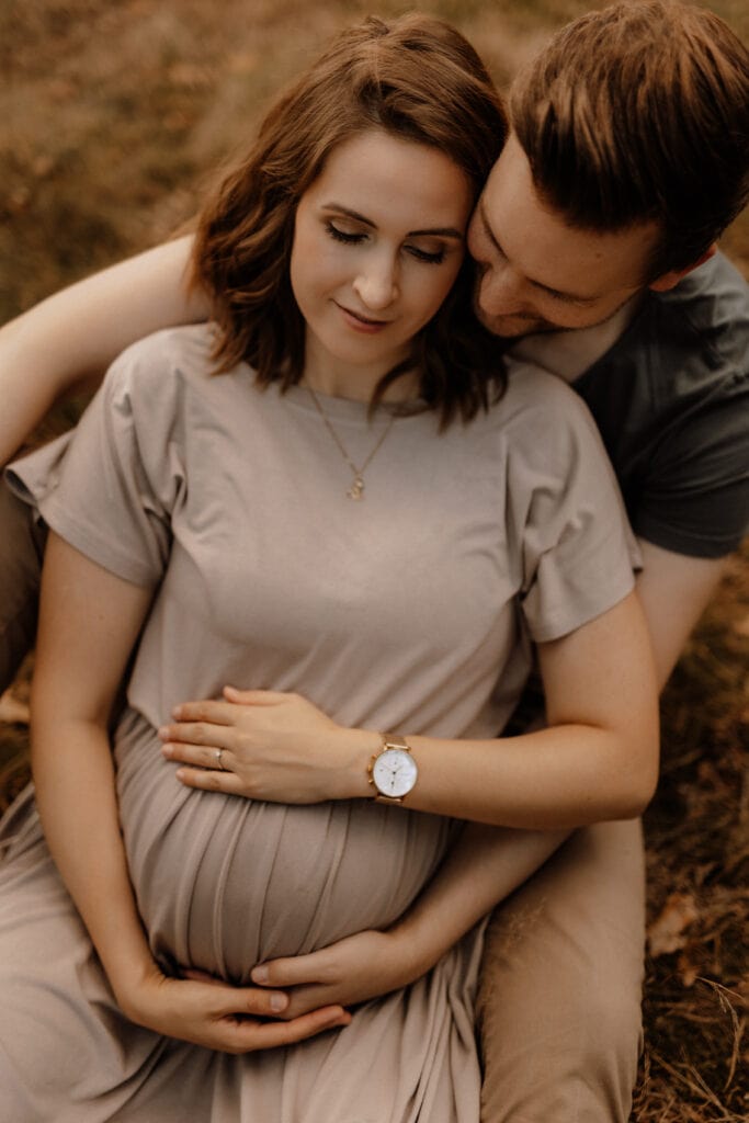 Eine schwangere Frau in einem hellgrauen Kleid sitzt auf dem Boden und hält mit beiden Händen ihren Babybauch. Dicht hinter ihr sitzt ein Mann, der seine Arme um sie geschlungen hat, seine Hände auf ihren ruhen lässt und liebevoll ihren Hals streichelt. Beide wirken zufrieden und friedlich.