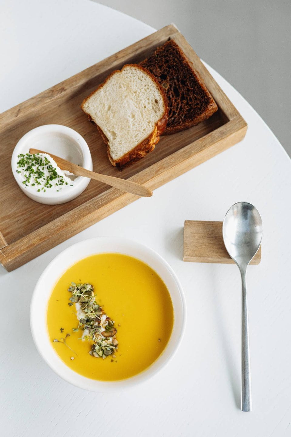 Auf einem weißen Tisch steht eine Schüssel gelbe Suppe, garniert mit Gemüse, neben einem silbernen Löffel. Daneben liegt ein Holztablett mit gerösteten und normalen Brotscheiben, einer kleinen Schüssel weißer Sahne und einem Holzlöffel.