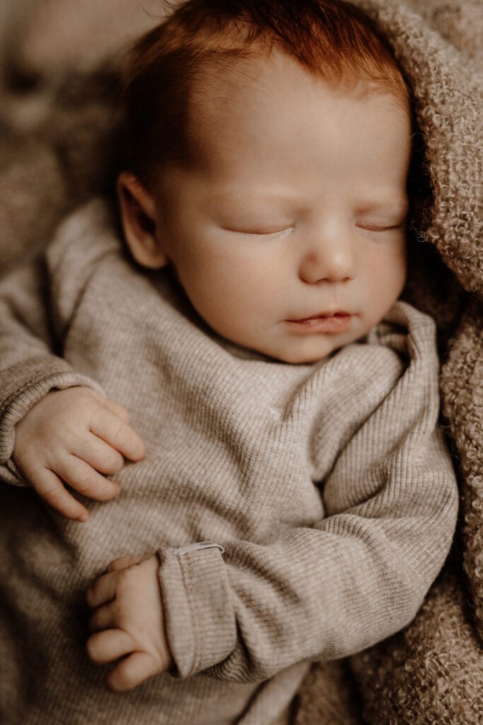 Eine Nahaufnahme eines Neugeborenen mit hellrotem Haar aus Havixbeck und Umgebung, das ein beiges langärmliges Outfit trägt und teilweise in eine weiche, strukturierte Decke gehüllt ist. Das Baby sieht friedlich und zufrieden aus, mit geschlossenen Augen und einem ruhigen Gesichtsausdruck.