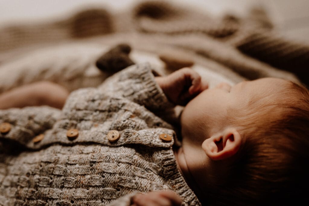 Ein Baby in einem Strickpullover liegt auf einer weichen, strukturierten Decke und fängt einen perfekten Moment für Neugeborenenfotografie ein. Mit einer Hand in der Nähe des Mundes und friedlich auf der Seite ruhend, wirkt die Szene warm und gemütlich im Kontrast zum groben, neutralen Muster der Decke darunter.