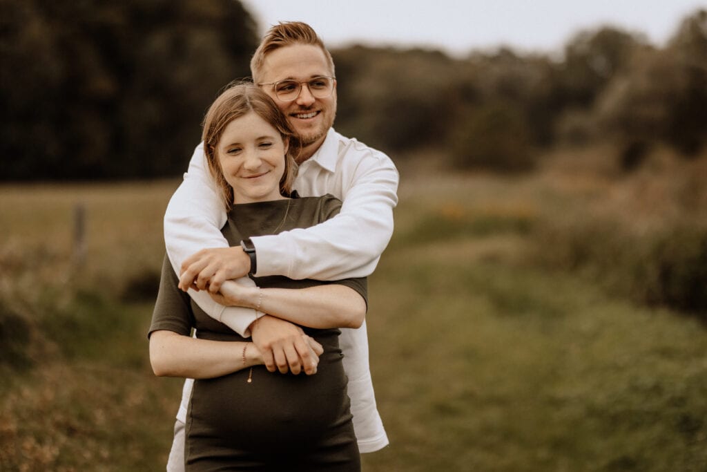 Ein Mann und eine Frau stehen auf einem Feld, wobei der Mann die Frau von hinten umarmt. Beide lächeln zufrieden. Die Frau, in ein grünes Kleid gekleidet, wiegt ihren Babybauch, während der Mann in einem weißen Hemd und mit Brille stolz strahlt. Der Hintergrund ist verschwommen mit Grün und Bäumen von Havixbeck und Umgebung.