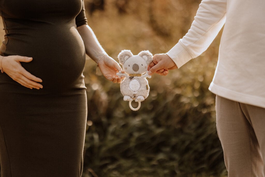 Eine schwangere Frau in einem dunklen Kleid hält ihren Bauch, während eine Person in einem weißen Oberteil und grauen Hosen neben ihr steht. Beide halten zwischen sich ein kleines Koala-Plüschtier. Der Hintergrund ist eine Außenkulisse mit viel Grün und sanftem Sonnenlicht, wunderschön eingefangen von einer Familienfotografin, die sich auf Babybauchfotografie in Havixbeck und Umgebung spezialisiert hat.