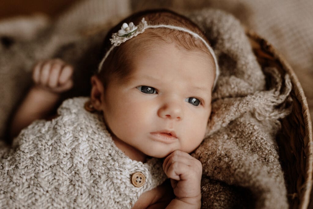 Ein Neugeborenes mit blauen Augen liegt auf einer strukturierten braunen Decke und trägt ein gestricktes cremefarbenes Outfit mit einem Holzknopf und ein Stirnband mit einer kleinen Blume. Während des Neugeborenen-Shootings schaut das Baby zur Seite und hat eine Hand in der Nähe seines Kinns.