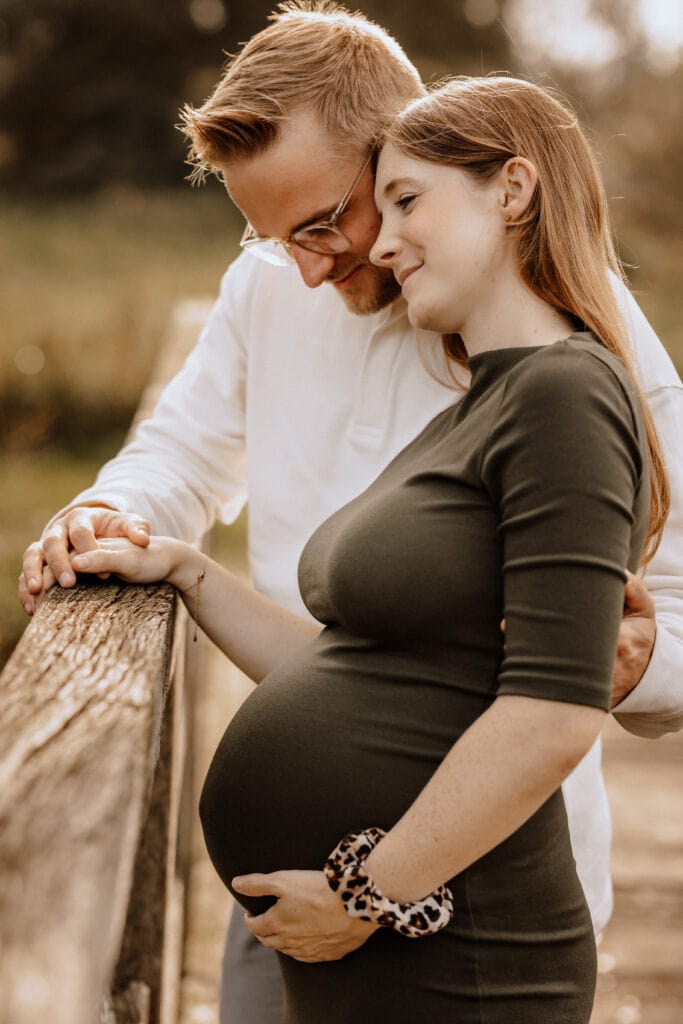 Eine schwangere Frau in einem grünen Kleid steht auf einer Holzbrücke und hält zärtlich ihren Babybauch. Sie wird von einem Mann in einem weißen Hemd begleitet, der sich nah an sie lehnt, beide lächeln und wirken liebevoll. Der Hintergrund ist sanft verschwommen und lässt eine ruhige Außenkulisse vermuten, die von ihrer Familienfotografin eingefangen wurde.