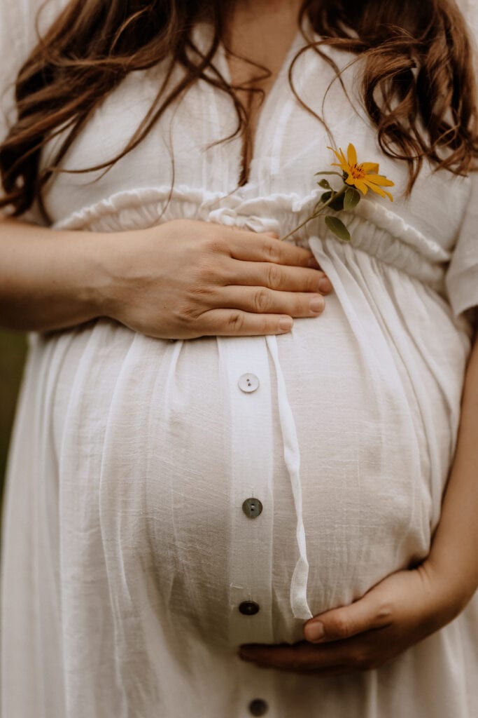 Eine schwangere Frau in Havixbeck und Umgebung trägt ein weißes Button-Down-Kleid und umarmt ihren Babybauch sanft mit beiden Händen. Ihr welliges, braunes Haar fällt über ihre Schultern und im oberen Knopfloch des Kleides steckt eine kleine gelbe Blume, perfekt für Babybauchfotografie.