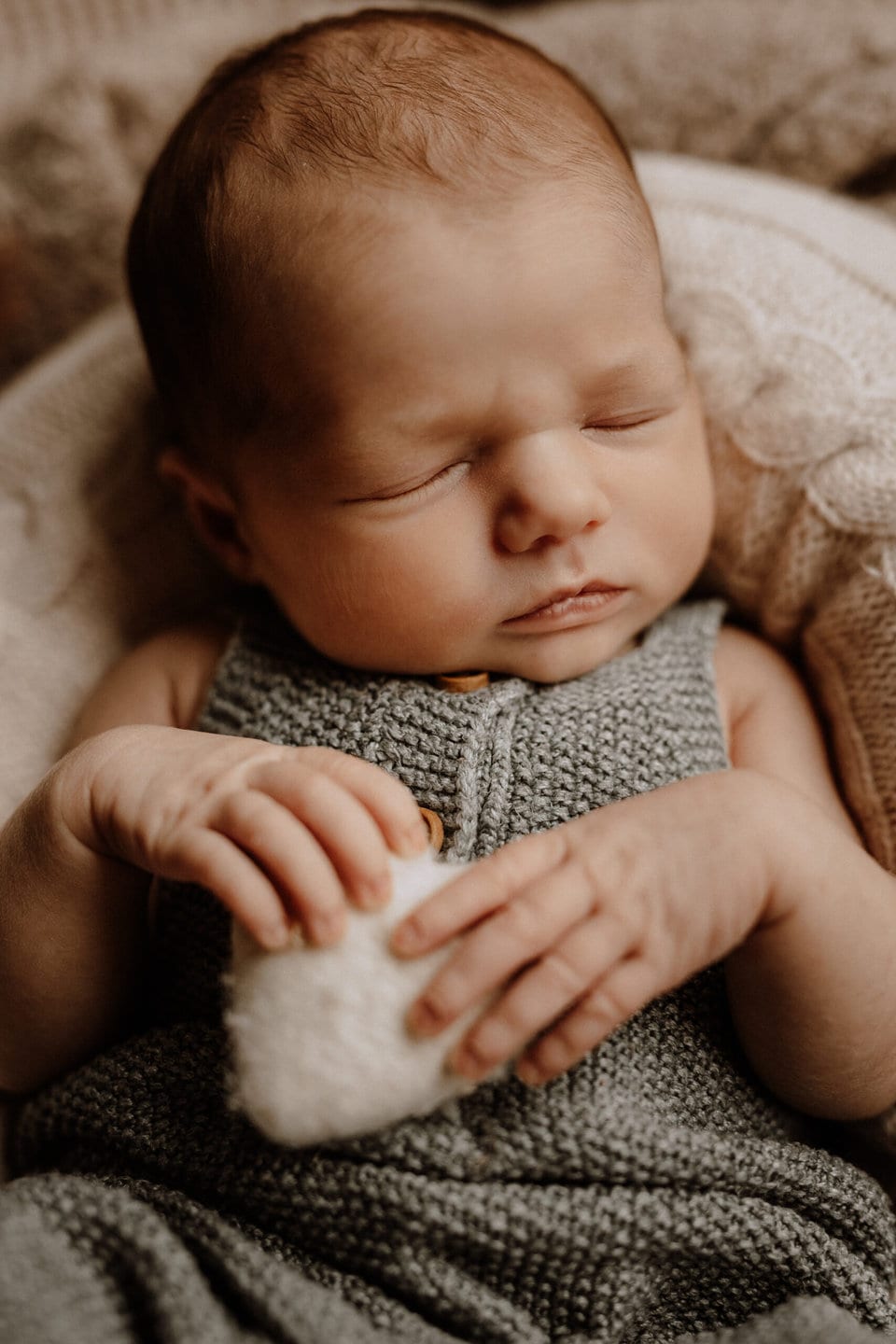 Ein Neugeborenes schläft friedlich in einem grünen Strickstrampler. Das Baby hält ein kleines, flauschiges weißes Stofftier an der Brust und ruht auf einer weichen Decke. Die zarten Gesichtszüge und der heitere Ausdruck des Babys vermitteln ein Gefühl von Ruhe und Unschuld, perfekt eingefangen in „Neugeborenenfotografie in Havixbeck und Umgebung“.