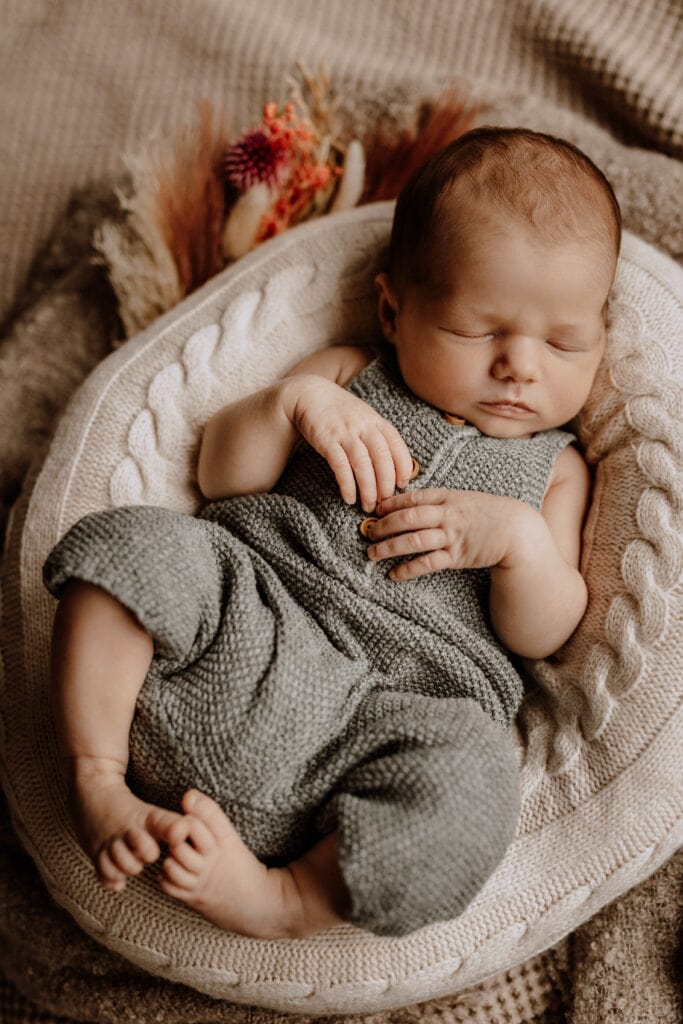Ein Neugeborenes schläft friedlich in einem gemütlichen, gepolsterten Korb. Das Baby trägt einen gestrickten grünen Overall und ist von weichen, gestrickten Decken und getrockneten Blumen umgeben. Die Atmosphäre ist warm und heiter mit gedämpften, erdigen Tönen. Die Essenz des Moments wird vom Newborn Fotografen aus Havixbeck und Umgebung eingefangen.