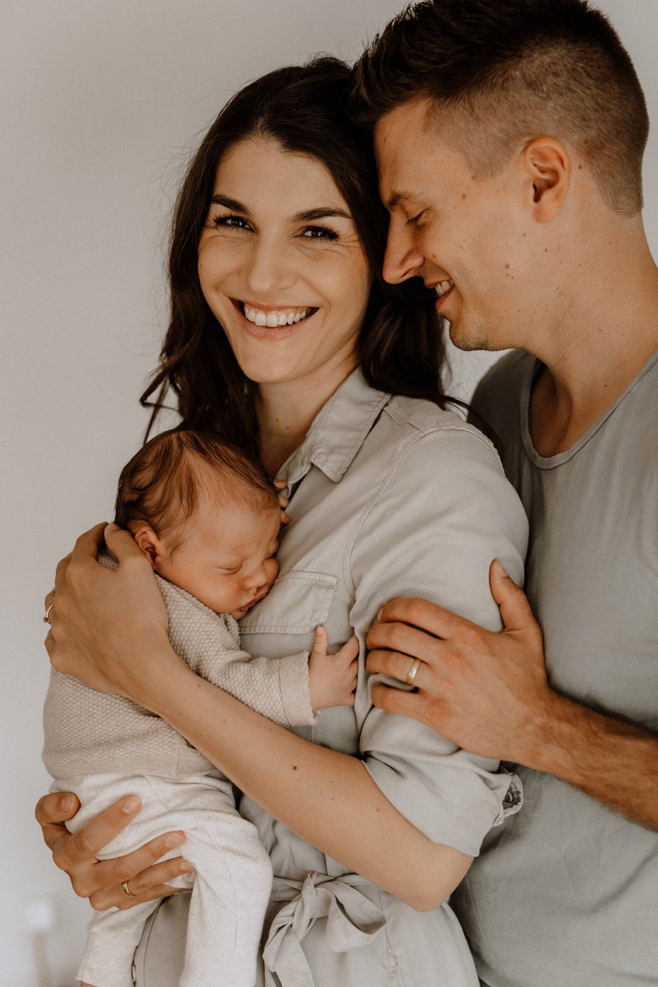 Eine Frau hält ein Neugeborenes an ihre Brust und lächelt warm in die Kamera. Ein Mann steht neben ihr und schaut das Baby mit freudigem Gesichtsausdruck an. Beide Erwachsenen sind leger gekleidet und der Moment wirkt zärtlich und liebevoll.