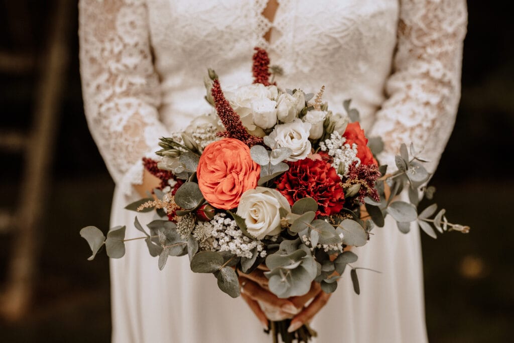 Eine Braut in einem Spitzenhochzeitskleid hält einen gemischten Blumenstrauß mit roten und weißen Rosen, roten Akzentblumen, Grünpflanzen und kleinen weißen Füllblumen. Die Hochzeitsfotografin aus Havixbeck und Umgebung fängt eine wunderschöne Stimmung ein.