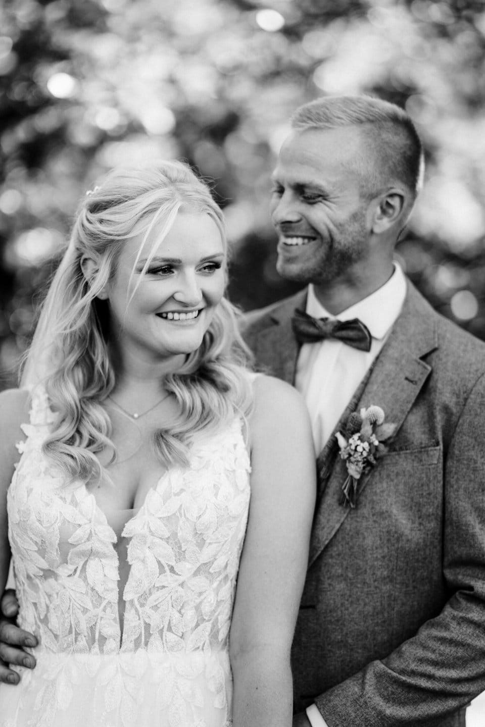 Ein Schwarzweißfoto eines lächelnden Paares an seinem Hochzeitstag, aufgenommen von einem Hochzeitsfotografen in Havixbeck und Umgebung. Die Frau, gekleidet in ein Spitzenhochzeitskleid, steht davor, während der Mann, in einem Anzug mit Boutonniere und Fliege, hinter ihr steht und sie mit freudigem Gesichtsausdruck ansieht. Sie sind im Freien.