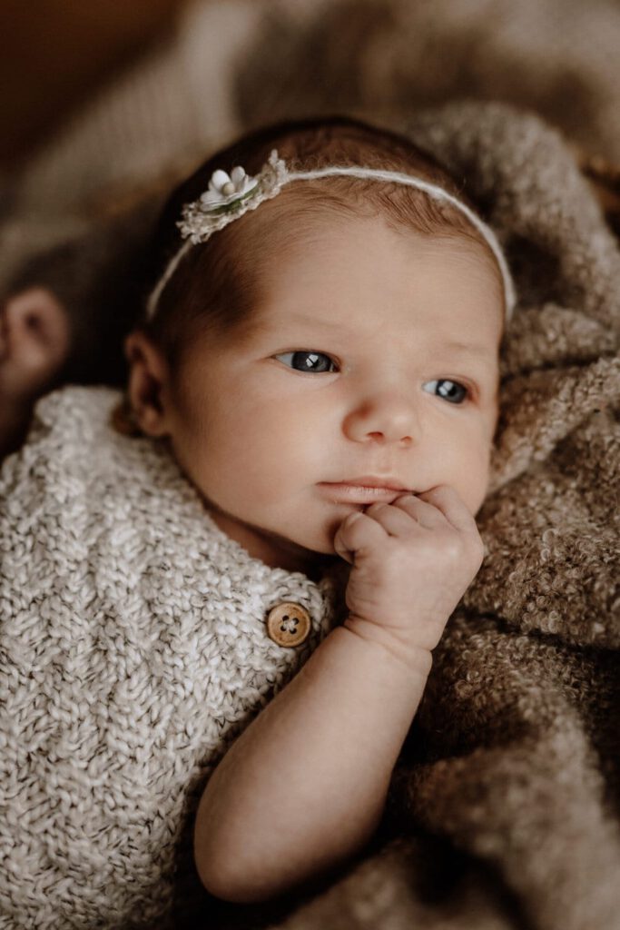 Eine Nahaufnahme eines Babys mit blauen Augen, das ein beiges Strickoutfit und ein Stirnband mit einer kleinen Blume trägt. Das Baby liegt auf einer weichen, strukturierten Decke und hat einen nachdenklichen Gesichtsausdruck, eine Hand unter dem Kinn – ein exquisites Beispiel für Neugeborenenfotografie im Münsterland