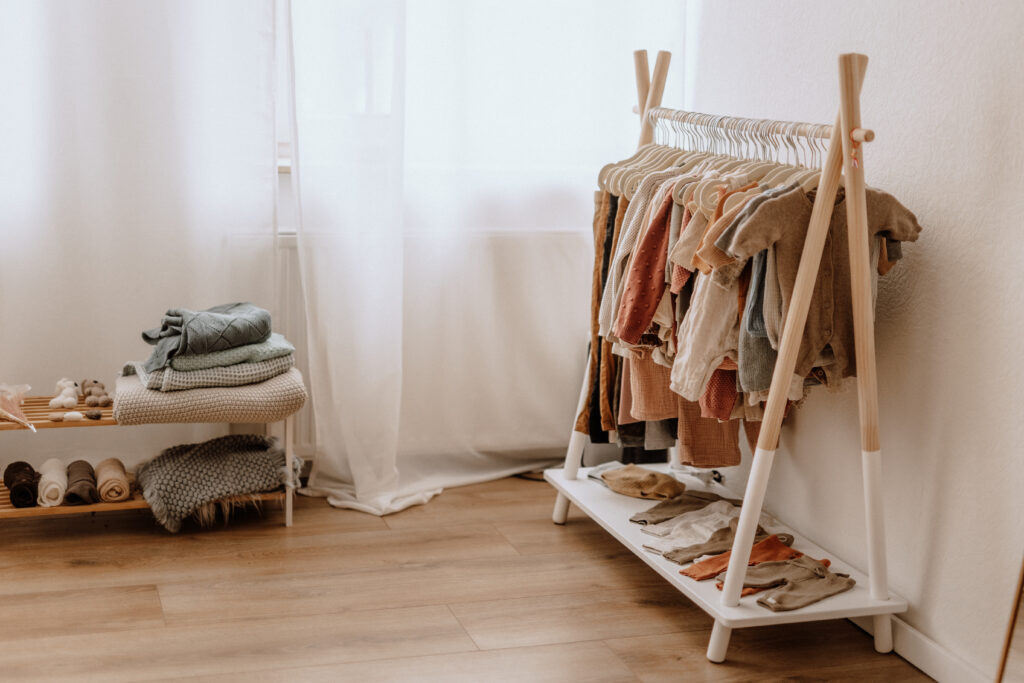 Ein kleiner kleiderständer präsentiert ordentlich aufgehängte kinderkleidung in neutralen tönen. daneben steht eine bank mit gefalteten decken und handtüchern. der raum hat helle holzböden und transparente weiße vorhänge.