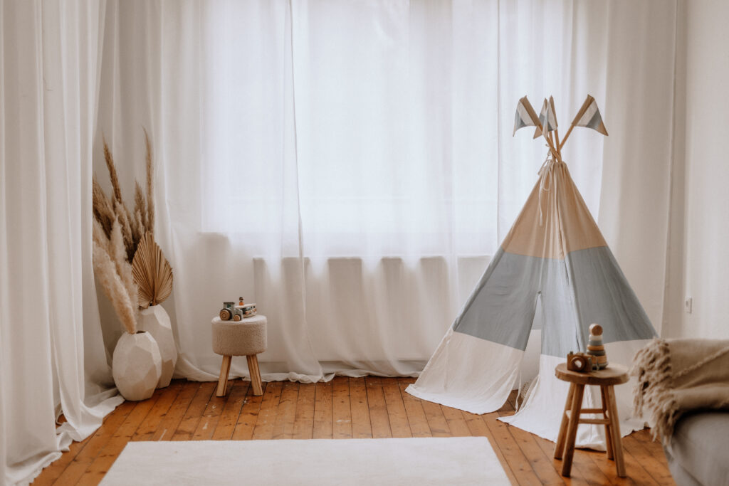 Ein gemütliches, minimalistisches kinderzimmer mit weißen vorhängen, einem beige blauen spielzelt, einem kleinen hocker, einem holzhocker mit spielzeug, einer großen vase mit getrockneten pflanzen und einem hellen teppich auf polierten holzdielen.