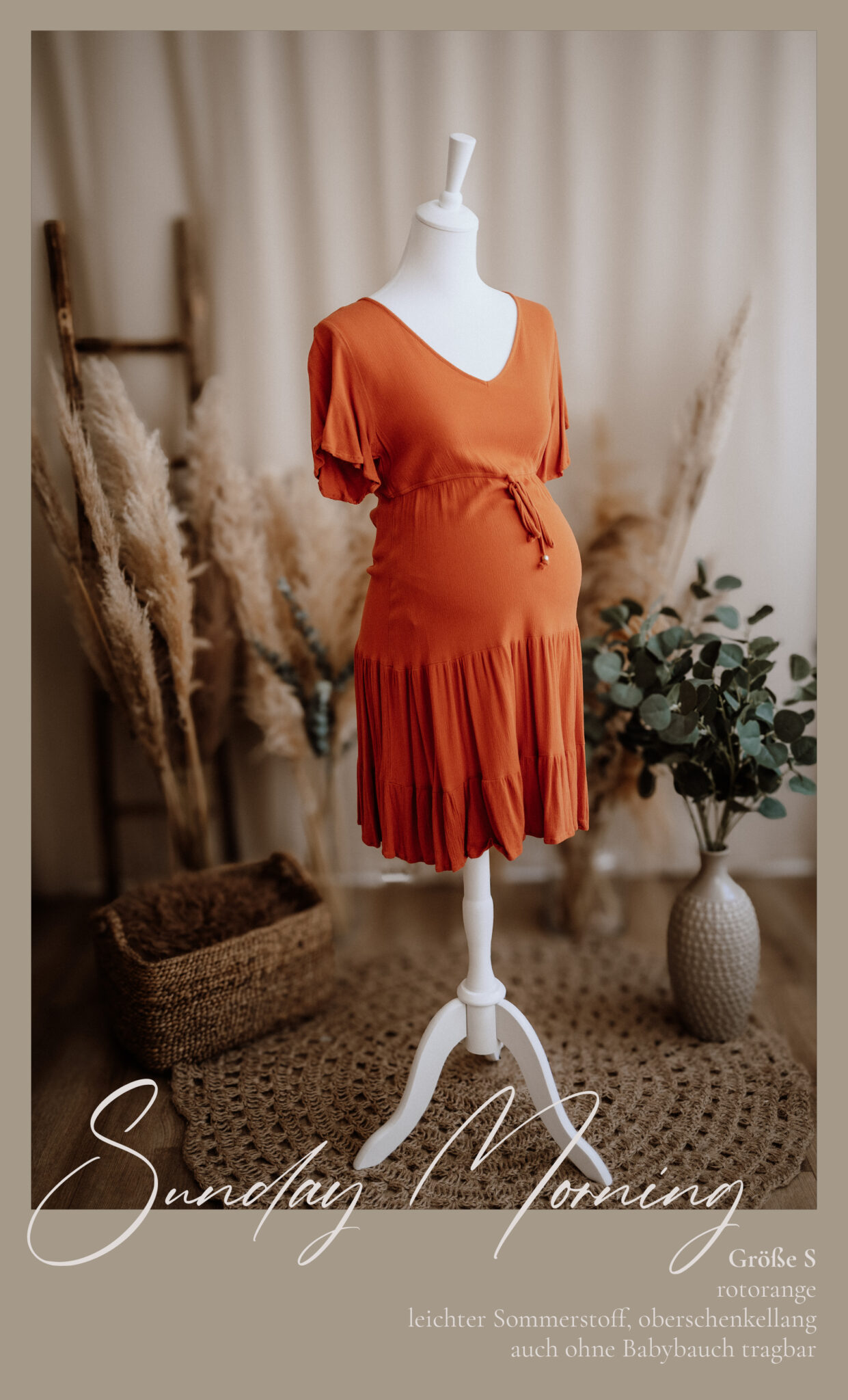 Ein rostorangefarbenes Umstandskleid mit Wickelmuster ist auf einer weißen Schaufensterpuppe ausgestellt. Die Umgebung besteht aus Pampasgras, einem Weidenkorb, einem runden Teppich und einer Vase, die einen gemütlichen, neutral getönten Hintergrund bilden.