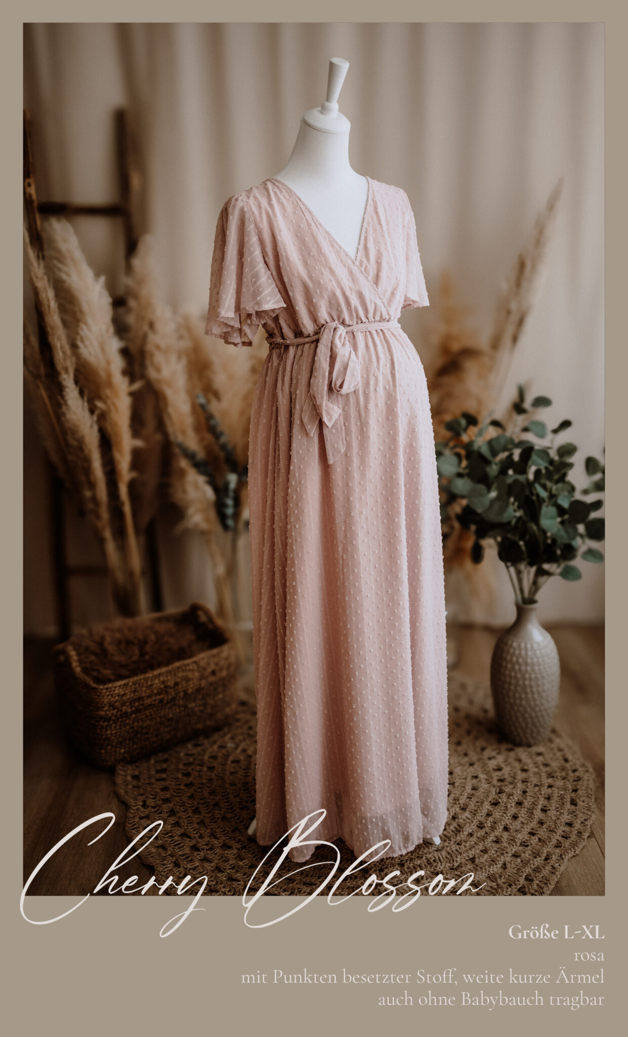 Ein blassrosa, kurzärmeliges, bodenlanges Umstandskleid mit Wickelmuster ist auf einer Schaufensterpuppe ausgestellt. Es steht auf einem gewebten Teppich mit dekorativen Körben, Pampasgras und Grünzeug in einem gemütlichen Raum. Der Text lautet: "Kirschblüte.".