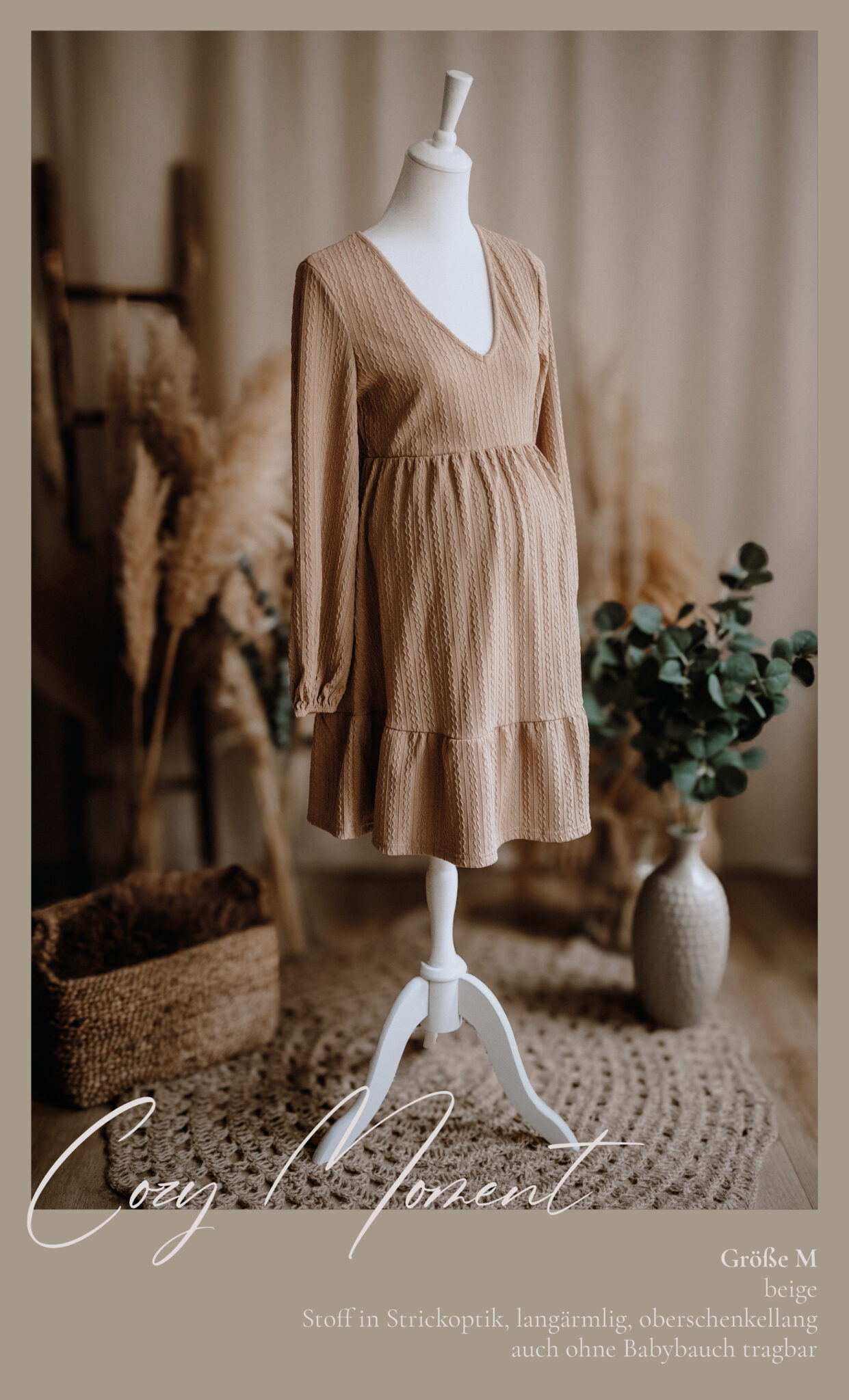 Ein beigefarbenes, langärmeliges, knielanges Umstandskleid mit V Ausschnitt, ausgestellt auf einer weißen Schaufensterpuppe. Der Hintergrund besteht aus Pampasgras, einem Weidenkorb und Eukalyptuszweigen, die eine gemütliche, natürliche Umgebung schaffen.